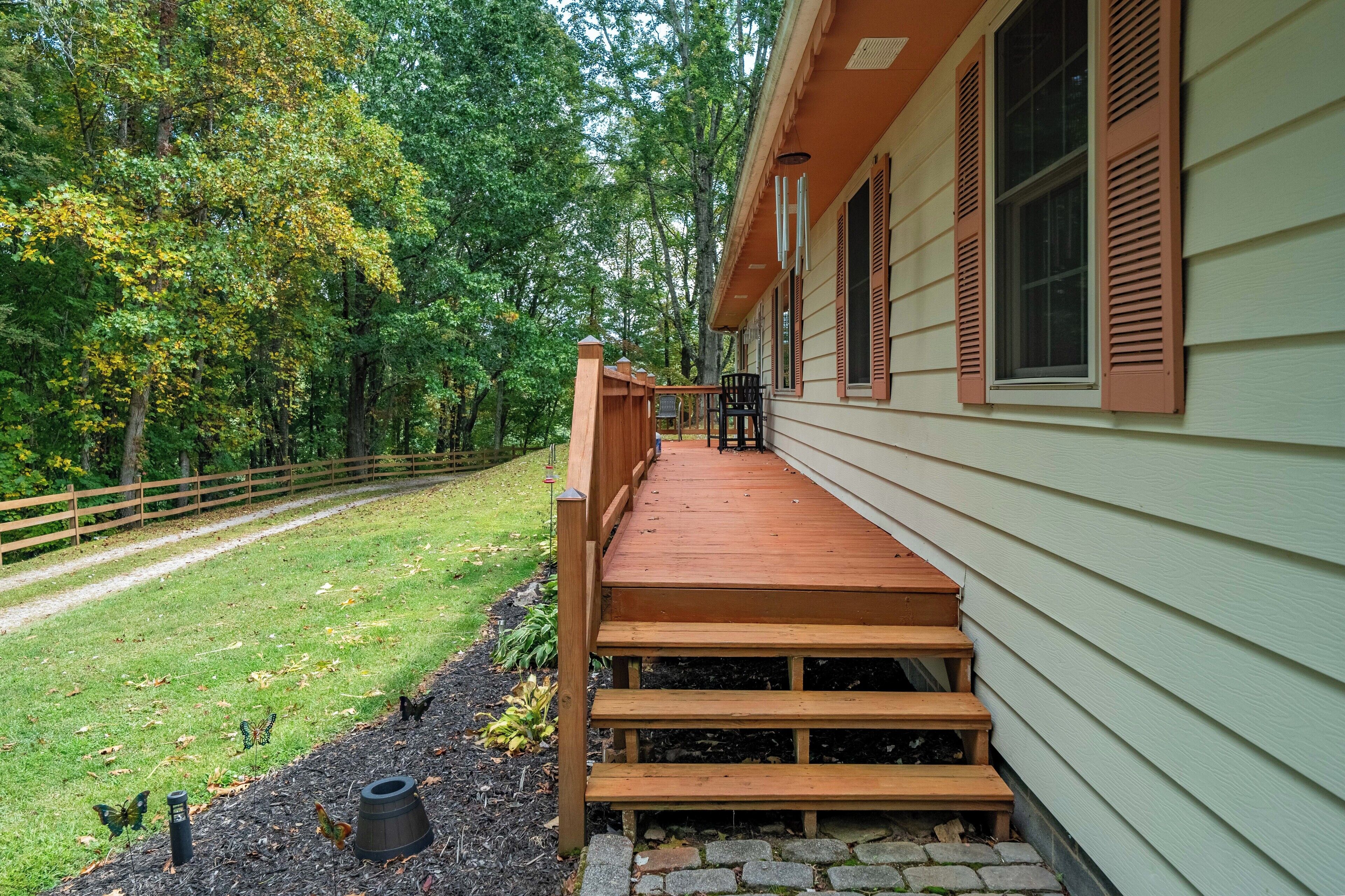 A wooden deck leading to a house with a porch, surrounded by a fenced yard and trees.