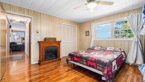 Ein Schlafzimmer mit Holzboden, einem Bett mit Decke, einem Kamin, einem Deckenventilator und einem Fenster mit Vorhängen.