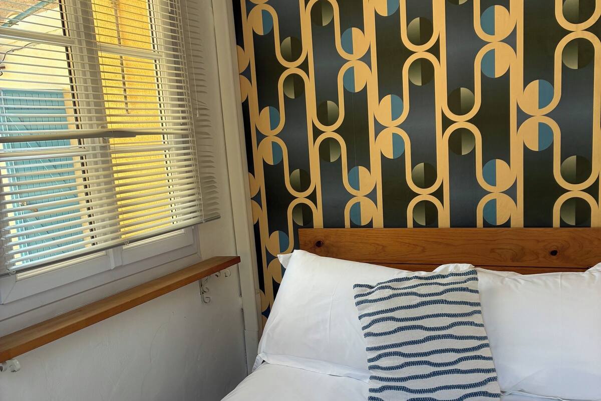 Un lit bien fait, avec du linge de lit blanc et un oreiller à motifs rayés. On y trouve des serviettes avec des inscriptions, et un mur orné d’un motif géométrique.