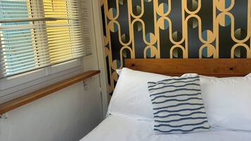 Ein ordentlich gemachtes Bett mit weißen Leinentüchern und einem Kissen mit Streifenmuster. Handtücher mit Aufdruck und eine Wand mit geometrischem Muster sind ebenfalls zu sehen.