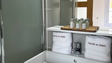 Un baño moderno con cabina de ducha de vidrio, un lavabo blanco y un espejo sobre él. Hay dos toallas blancas con el texto "Escale au Soleil" y una bandeja con artículos de aseo en el borde del lavabo.