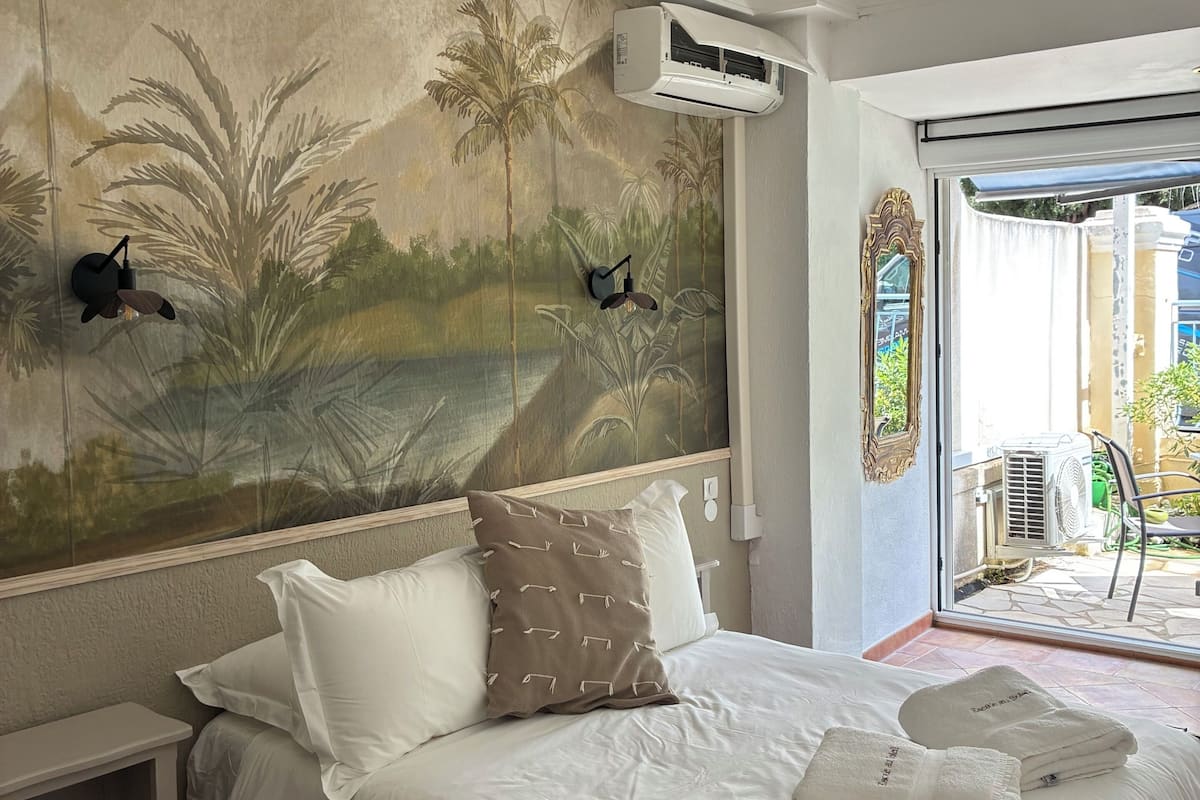 Une chambre à coucher avec un lit, des oreillers blancs, une fresque murale, un appareil de climatisation et une vue à travers une porte vitrée.