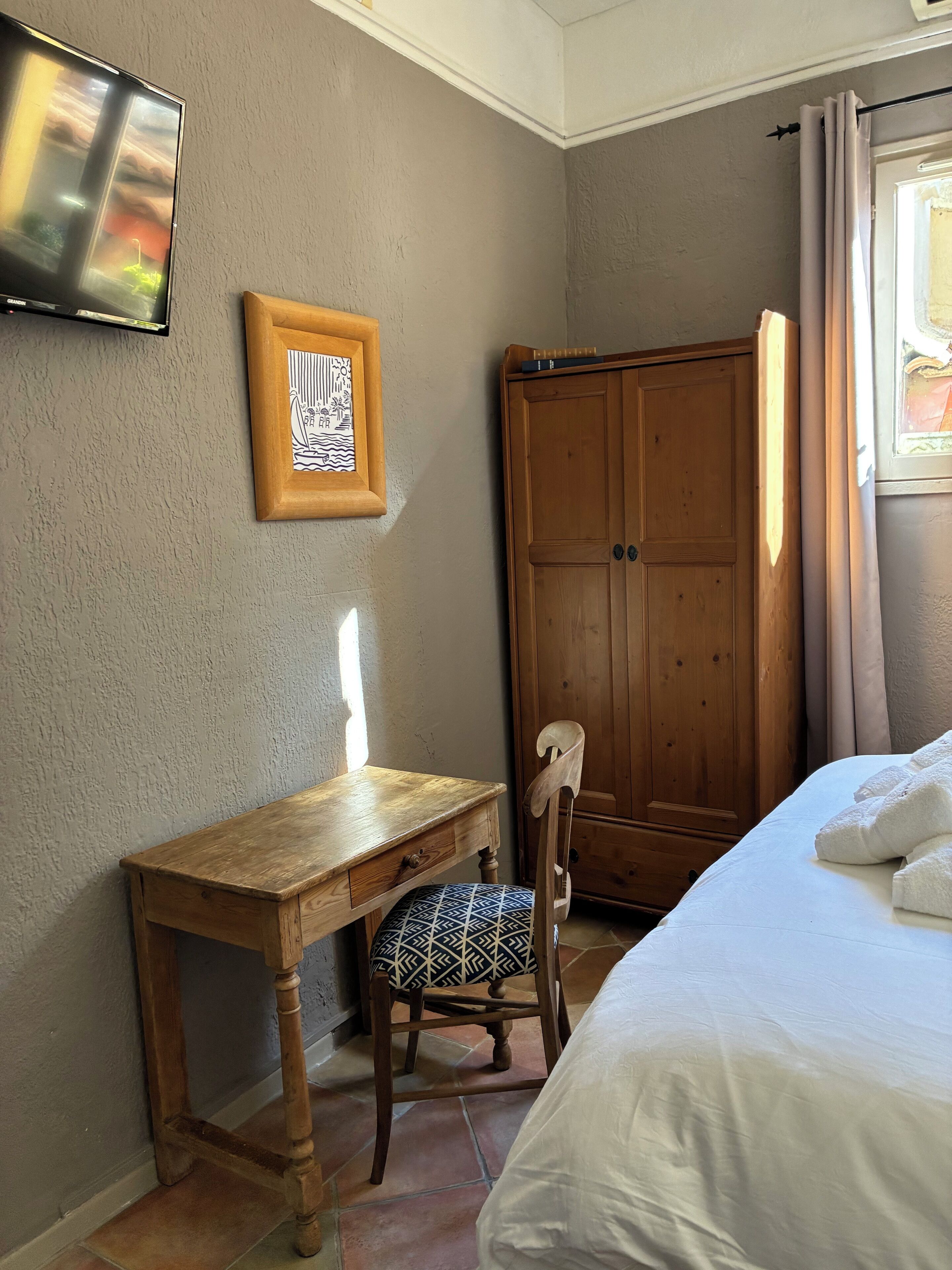 Ein Schlafzimmer mit einem Holztisch, einem Stuhl, einem Bett mit weißer Bettwäsche, einem Kleiderschrank und einem an der Wand montierten Fernseher.