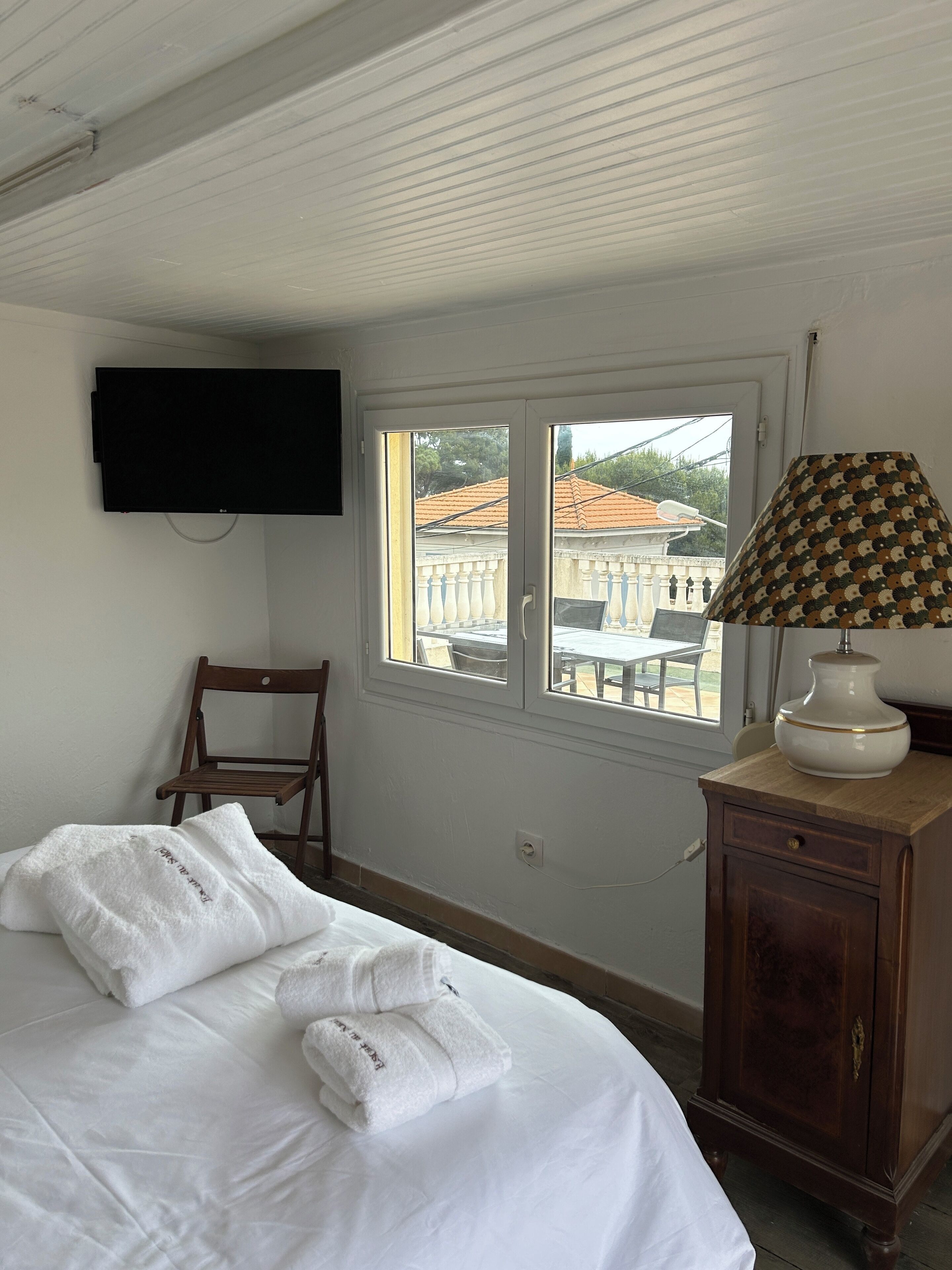 A hotel room with a bed, a television mounted on the wall, a window with a view of buildings, and a wooden cabinet.