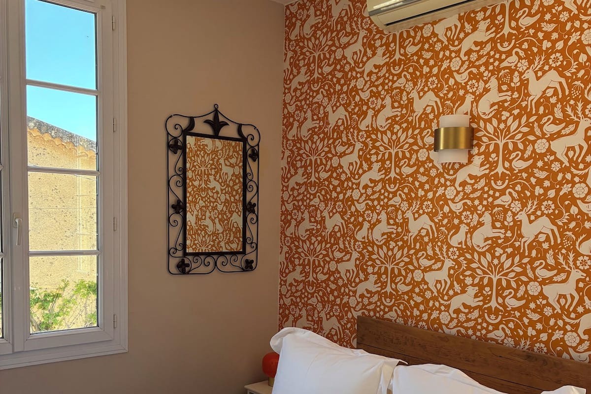 Un lit bien fait, avec du linge de lit blanc et une serviette, un climatiseur fixé au mur, une fenêtre donnant sur l’extérieur et un miroir décoratif.