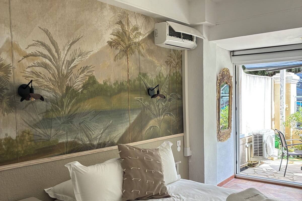 Une chambre à coucher avec un lit, des oreillers blancs, une fresque murale décorative, un appareil de climatisation et une vue à travers une porte vitrée.
