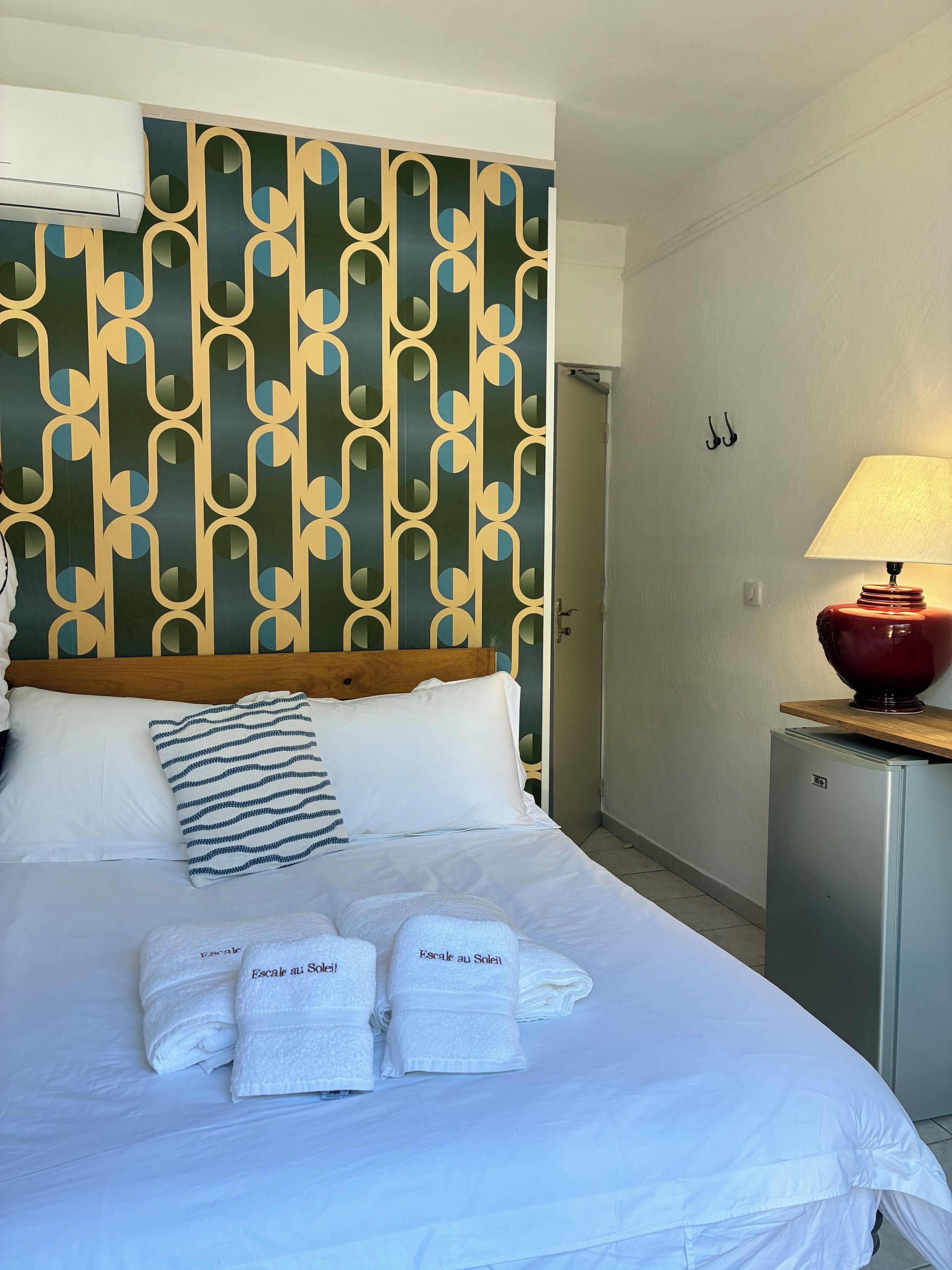 A hotel room with a patterned headboard, a bed with white linens, two towels with text, a red lamp, and a small refrigerator.