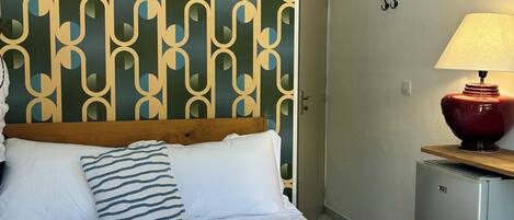 A hotel room with a patterned headboard, a bed with white linens, two towels with text, a red lamp, and a small refrigerator.