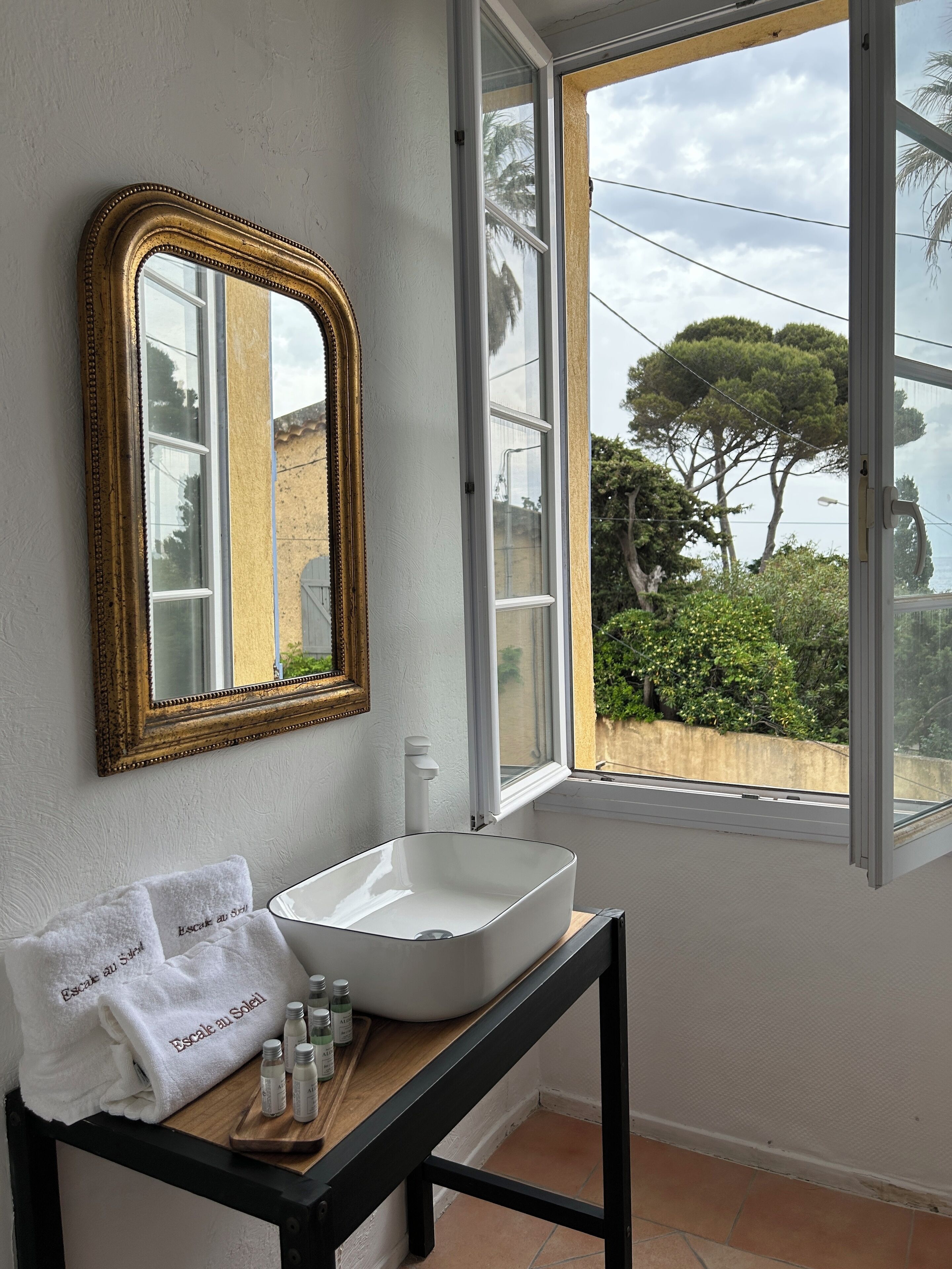 A bathroom with a white sink, a wooden vanity, a gold-framed mirror, and a window offering a view of trees and buildings.