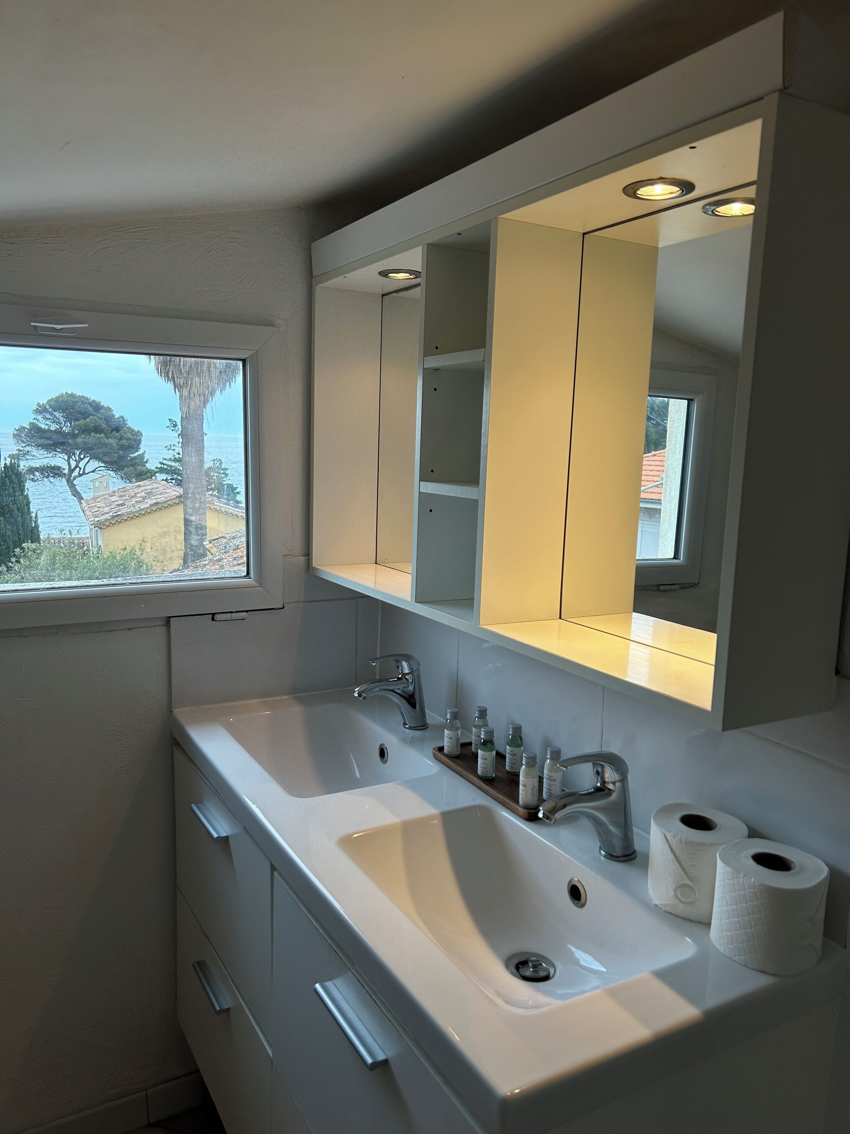 Ein Badezimmer mit großem Spiegel, zwei Waschbecken und einem Fenster mit Blick auf das Meer und Bäume.