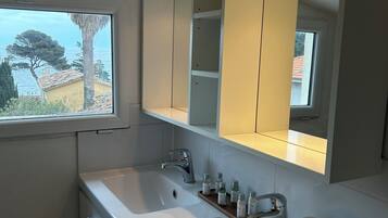 Ein Badezimmer mit großem Spiegel, zwei Waschbecken und einem Fenster mit Blick auf das Meer und Bäume.