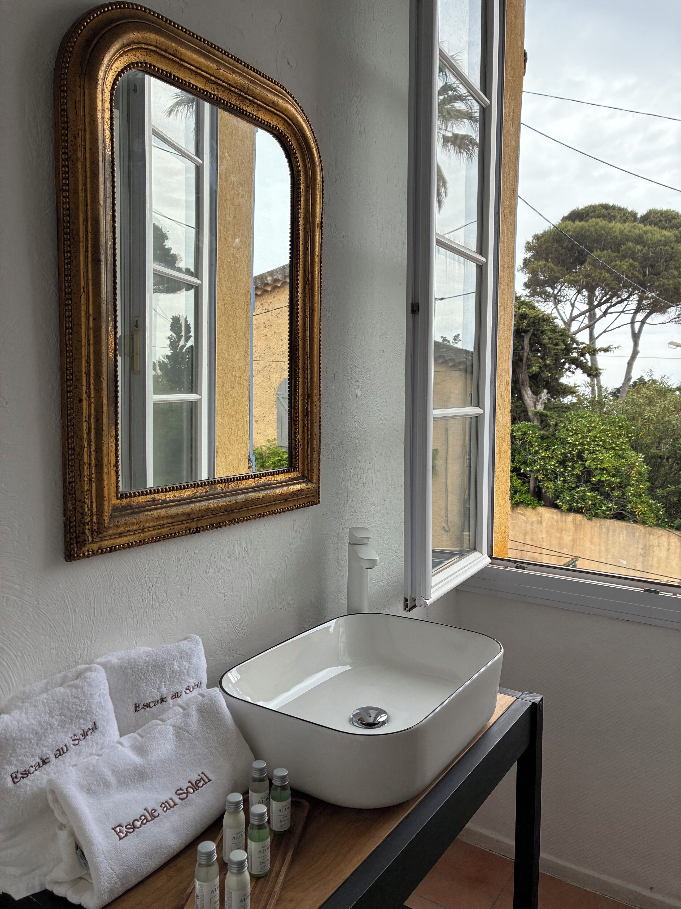 Une salle de bain avec un lavabo blanc, un miroir encadré d’or et une fenêtre donnant sur des arbres et des immeubles.