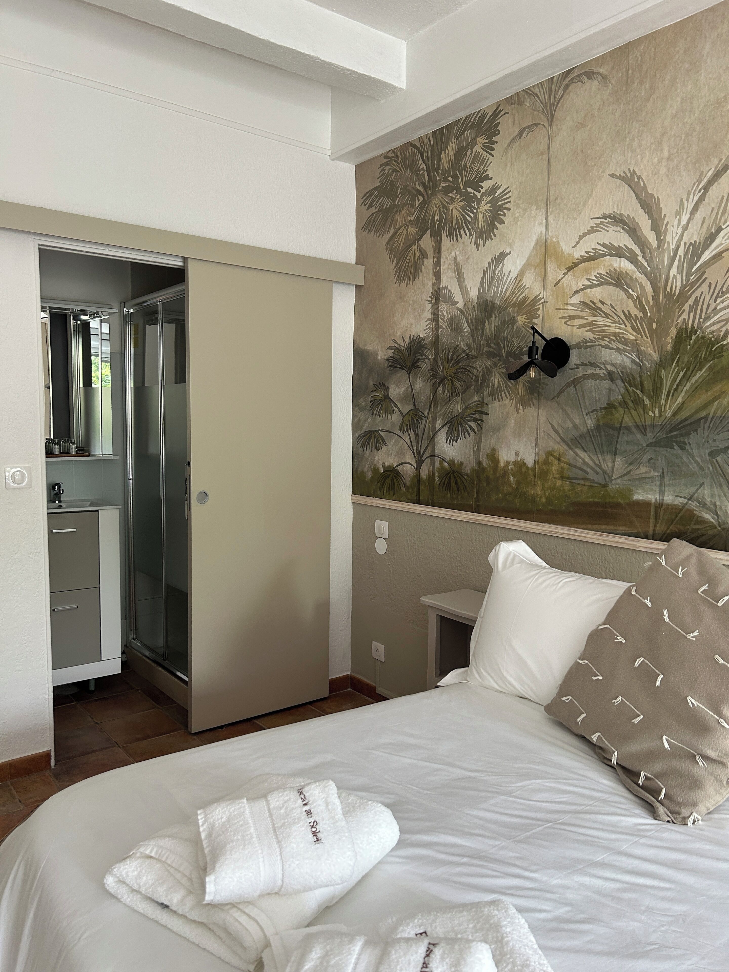 A hotel room with a bed, a painting of palm trees, a nightlight, and a bathroom with a shower and a mirror.