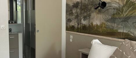 A hotel room with a bed, a painting of palm trees, a nightlight, and a bathroom with a shower and a mirror.