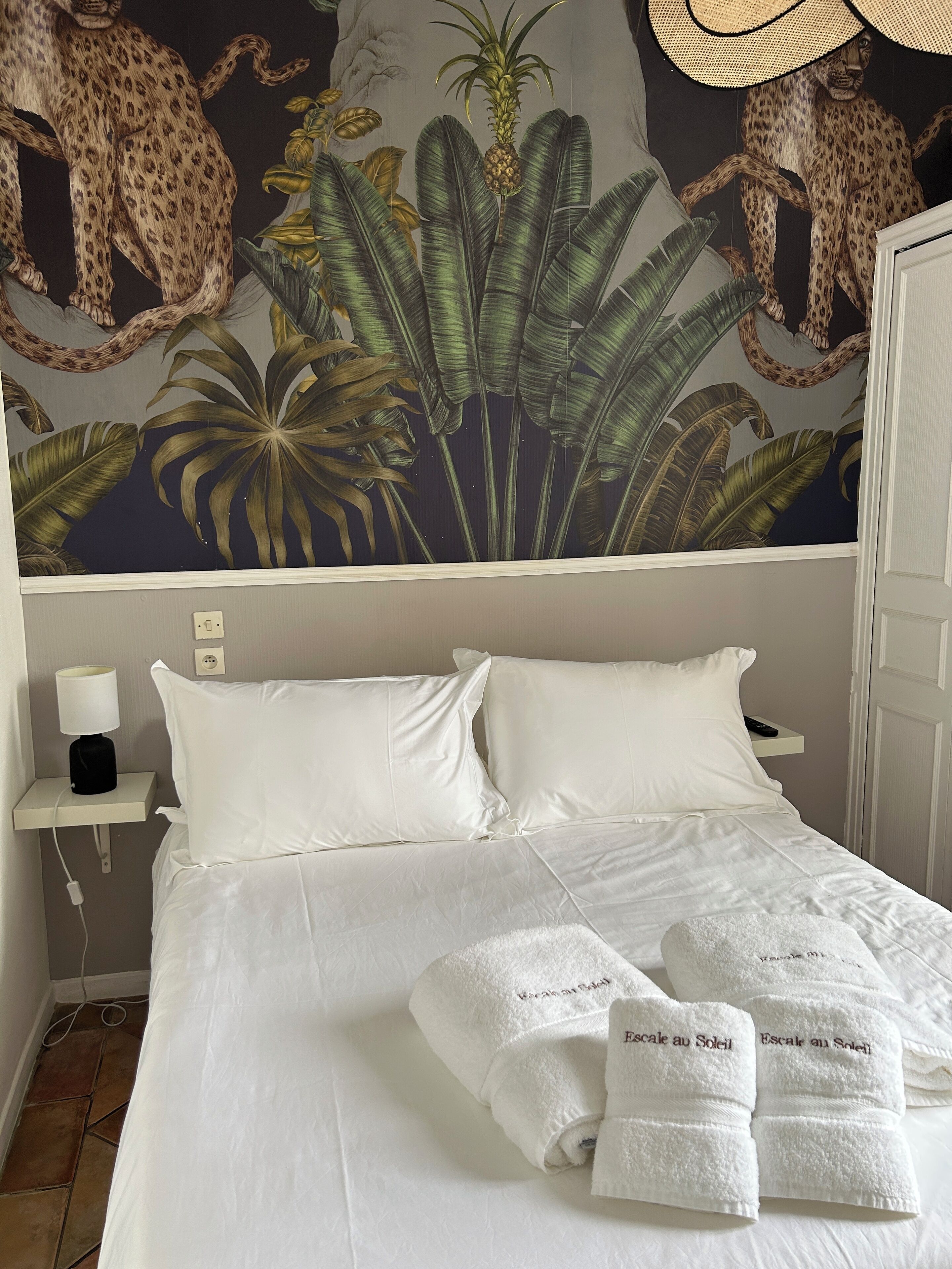 A neatly made bed with white linens and two pillows, a bedside table with a lamp, and a wall mural featuring a leopard and tropical plants.