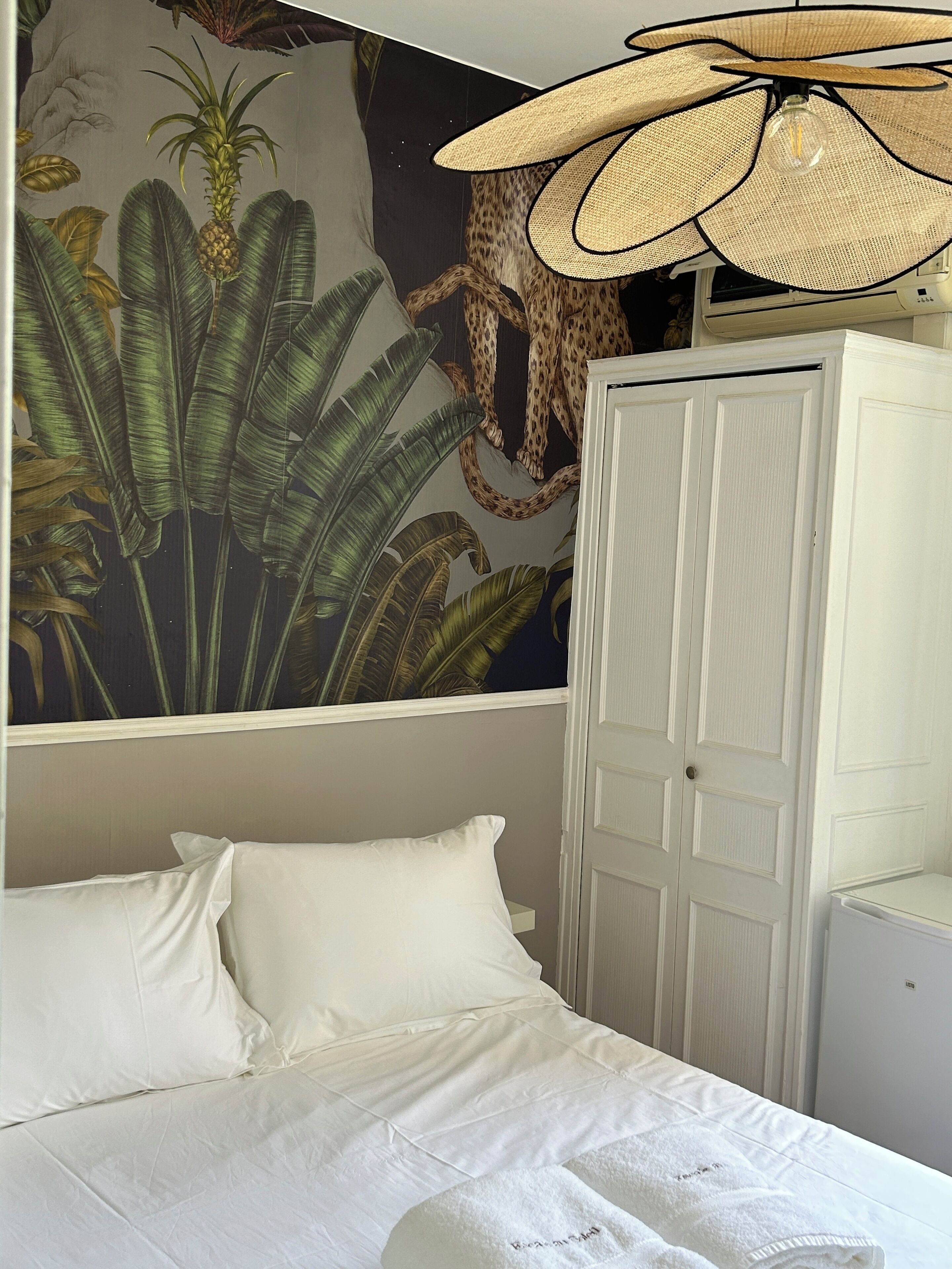 A bedroom with a bed, white pillows, a white wardrobe, and a mural of a leopard and foliage on the wall.