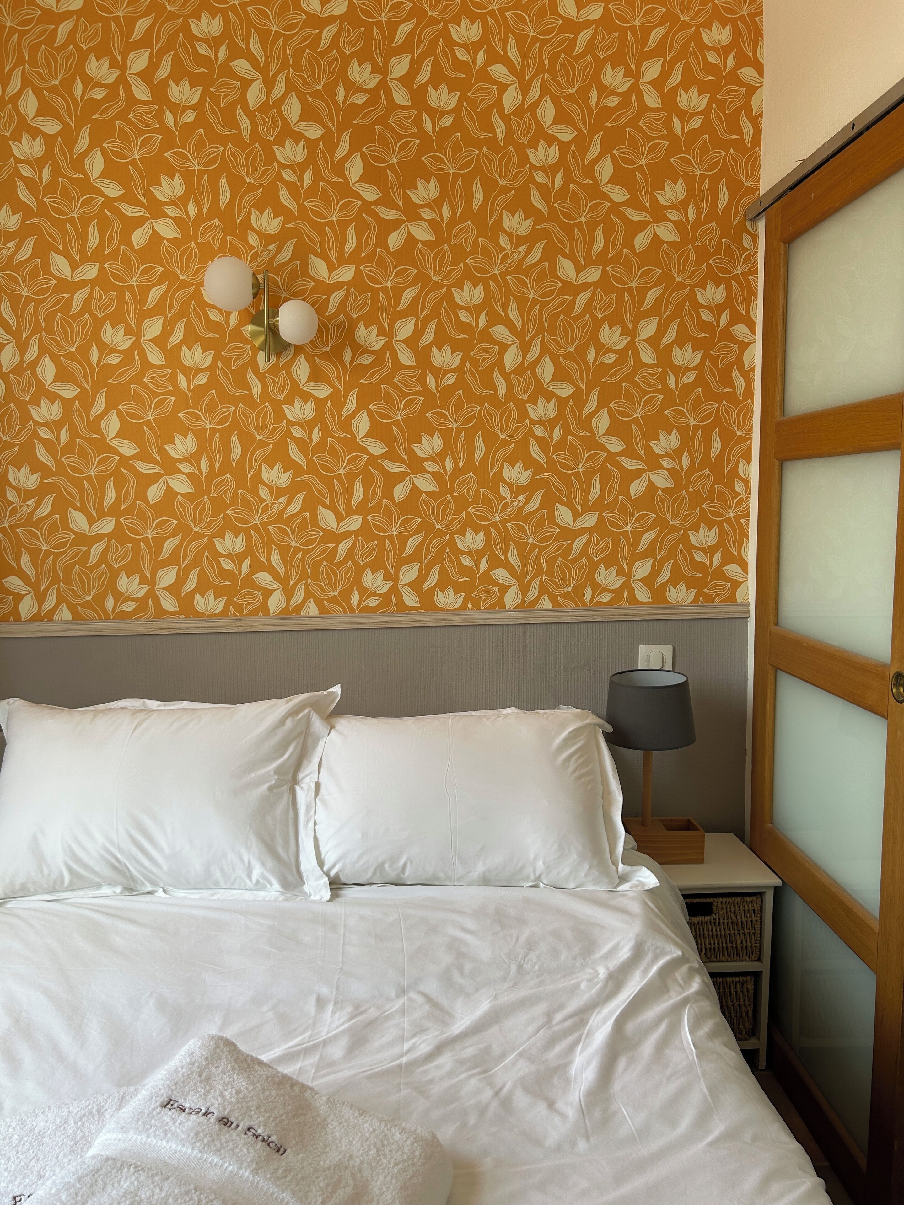 Una habitación de hotel con cama, ropa de cama blanca, una mesita de noche con lámpara y un papel tapiz con diseño floral.