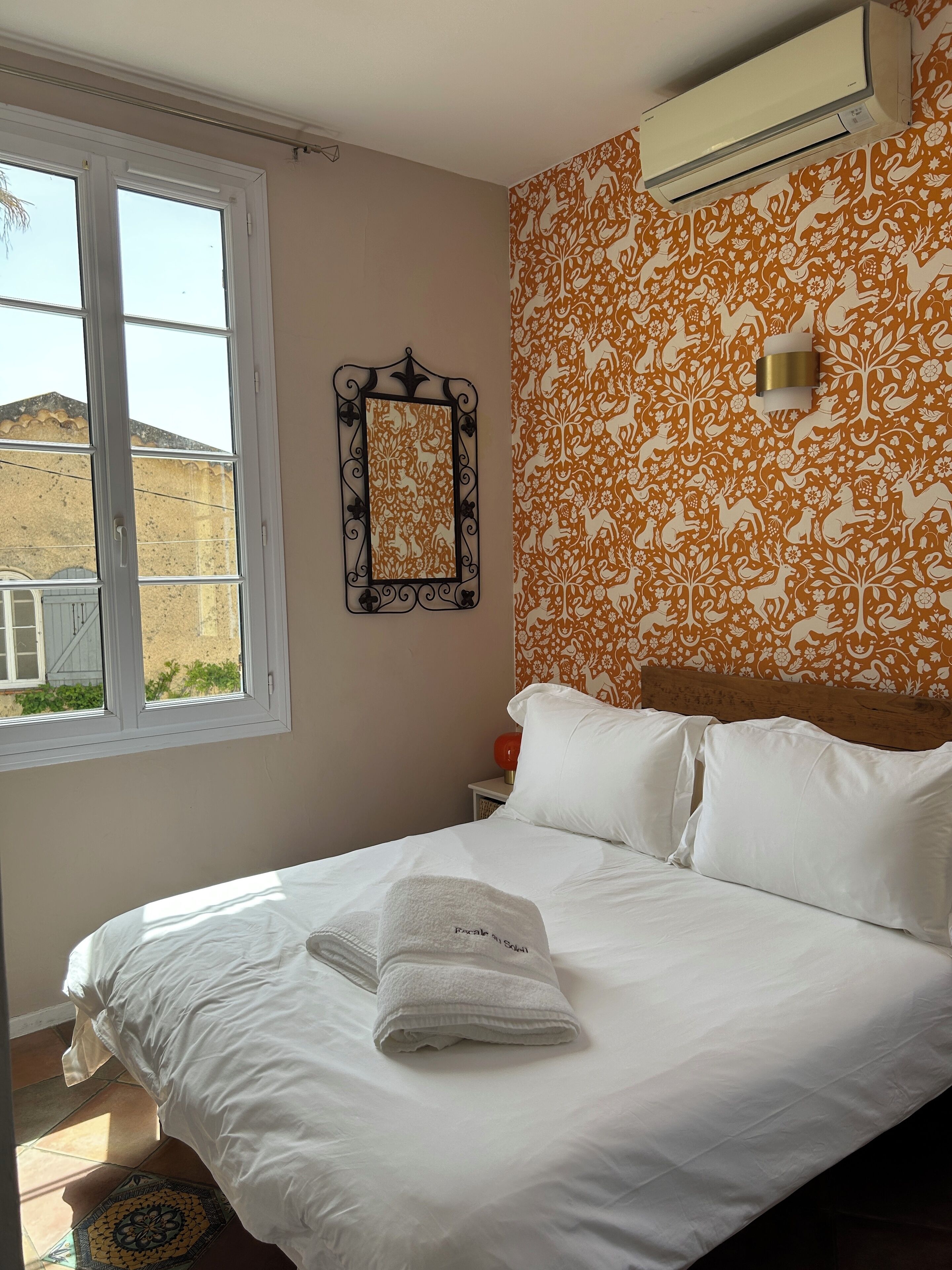 A bedroom with a bed, white bedding, a window, an air conditioner, a patterned wall, and a small table with a red lamp.