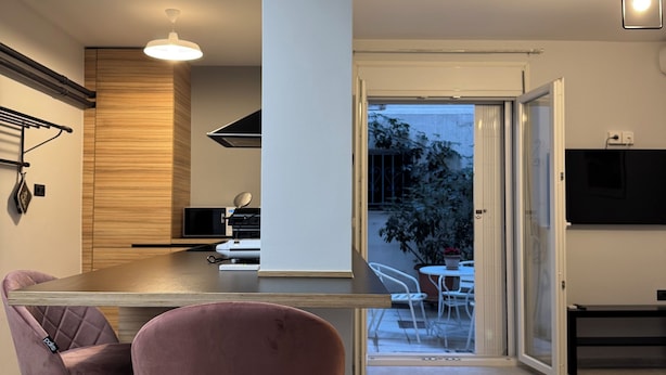 A modern kitchen with a central island, two pink bar stools, a dining area with a table and chairs, and a sliding glass door leading to an outdoor patio.