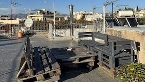 A rooftop terrace with a wooden bench, a potted plant, and a view of buildings in the background.