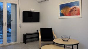 A modern living room with a flat-screen TV, a painting, a round table with a bowl, a black chair, and a sliding glass door leading to an outdoor area.