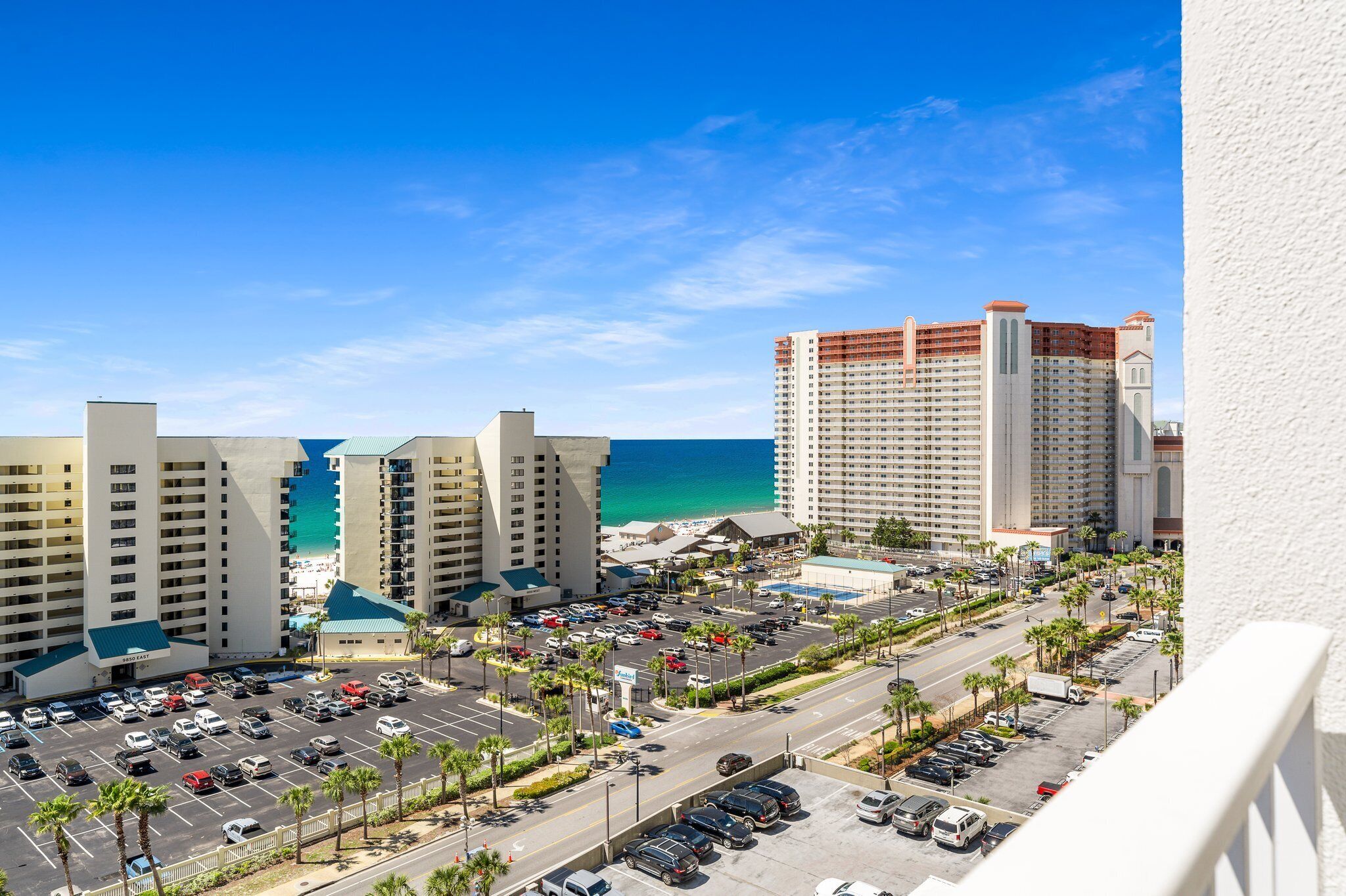 A high-rise hotel with a view of the ocean, a parking lot, and a road with palm trees.