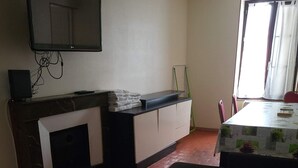 A room with a fireplace, a TV mounted on the wall, a dining table with chairs, and a window with curtains. - SPACIOUS 3-BEDROOM APT (APT N°15) (Roanne)