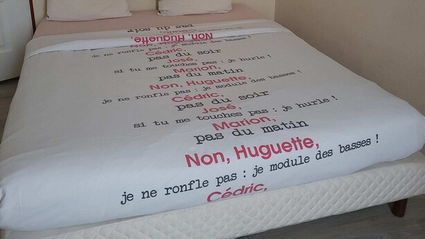 A bed with a white mattress and a quilted cover. There are two pillows on the bed, one striped and one plain. The bed is positioned against a plain wall. - SPACIOUS 3-BEDROOM APT (APT N°15) (Roanne)