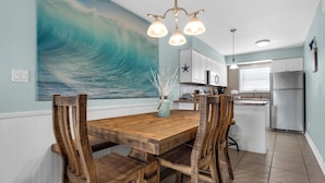 Un coin repas avec une table et des chaises en bois, une cuisine en arriÚre-plan et un grand tableau représentant une vague accroché au mur.