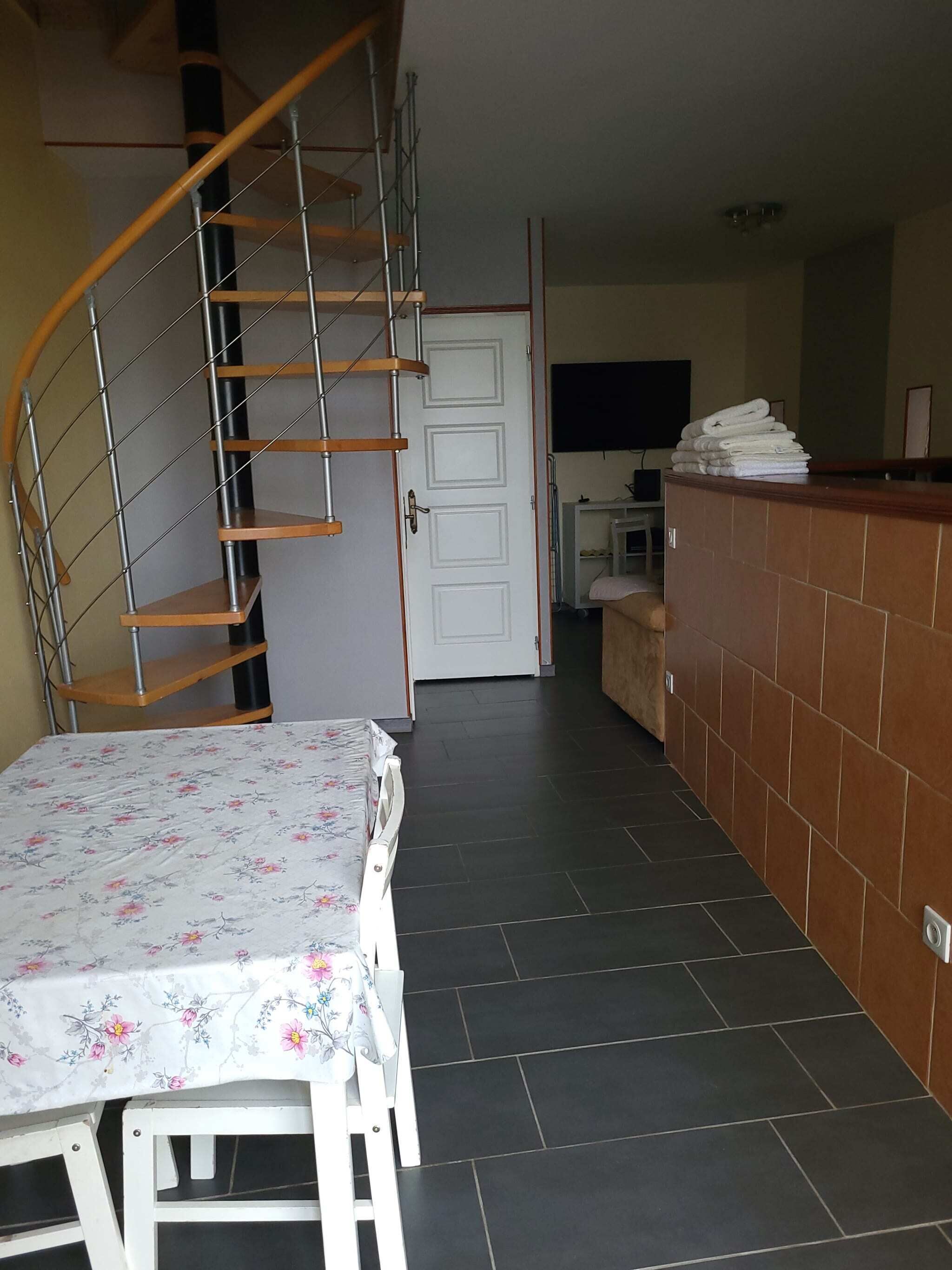 A room with a spiral staircase, a bed with a floral bedspread, a tiled floor, a kitchen area, and a counter with towels.