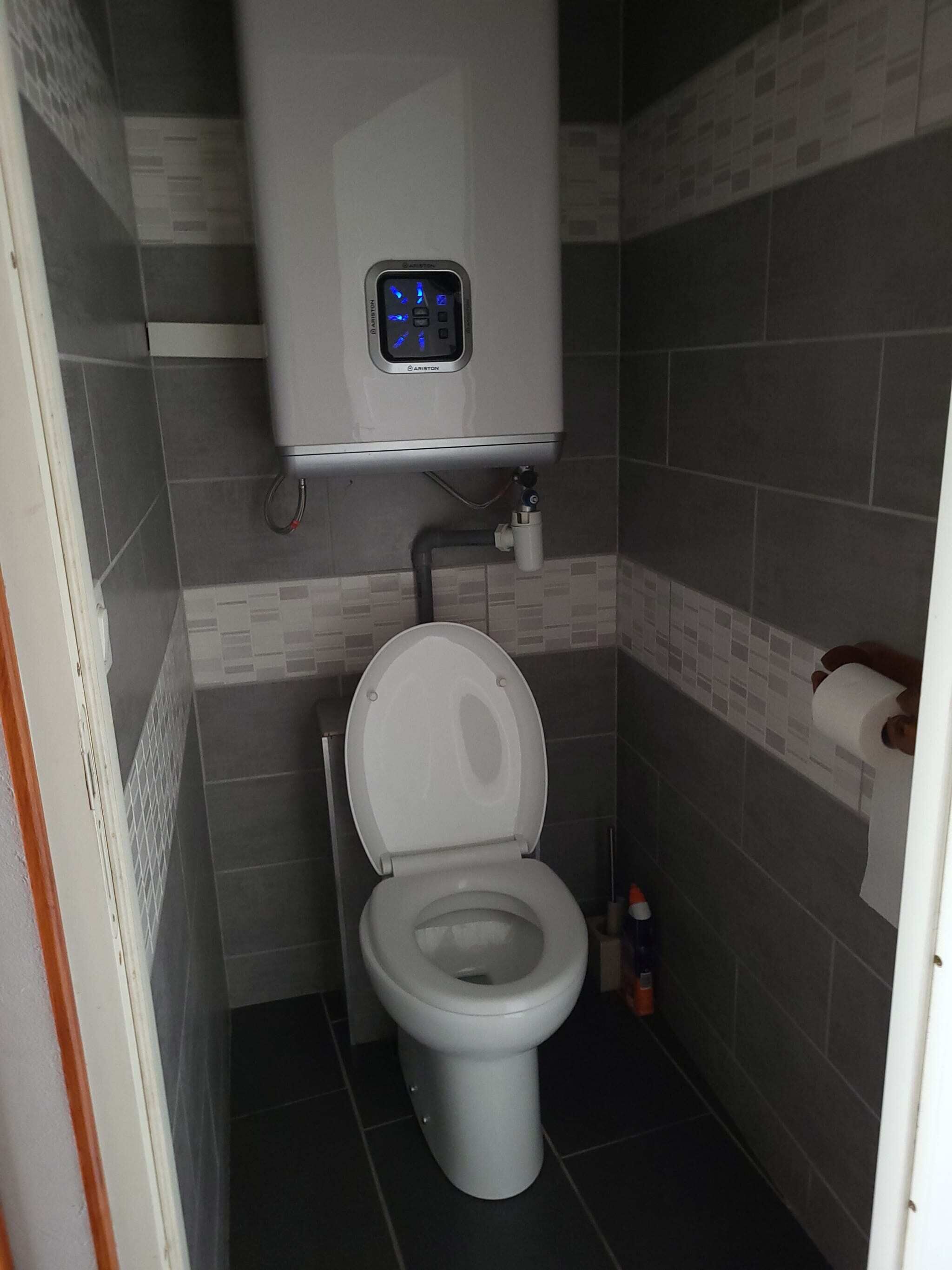 Une petite salle de bain moderne, équipée de toilettes, d’un réservoir de chasse d’eau fixé au mur et d’un porte-rouleau de papier toilette.