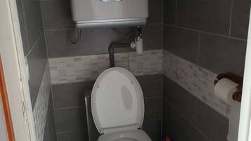 Une petite salle de bain moderne, équipée de toilettes, d’un réservoir de chasse d’eau fixé au mur et d’un porte-rouleau de papier toilette.