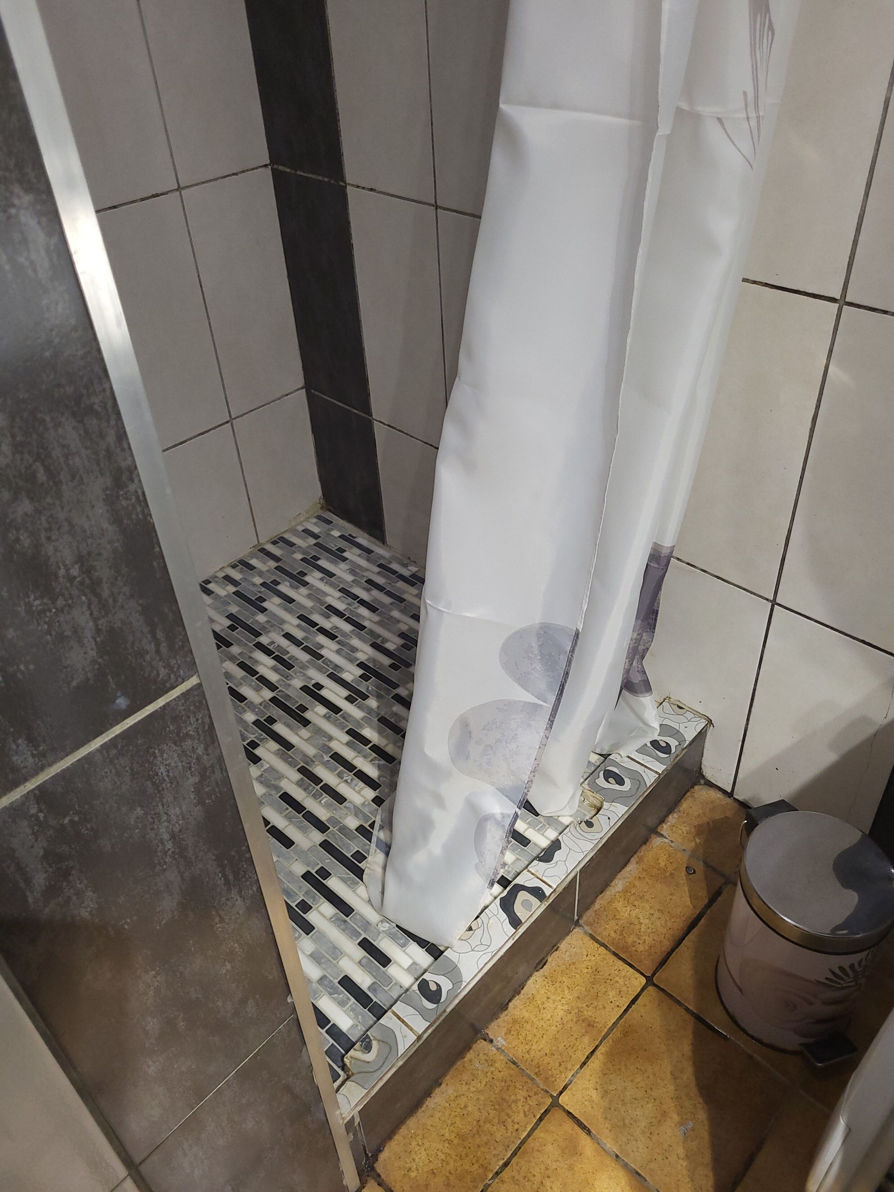 En dusch med ett mönstrat golv, en vit duschdraperi och en papperskorg.