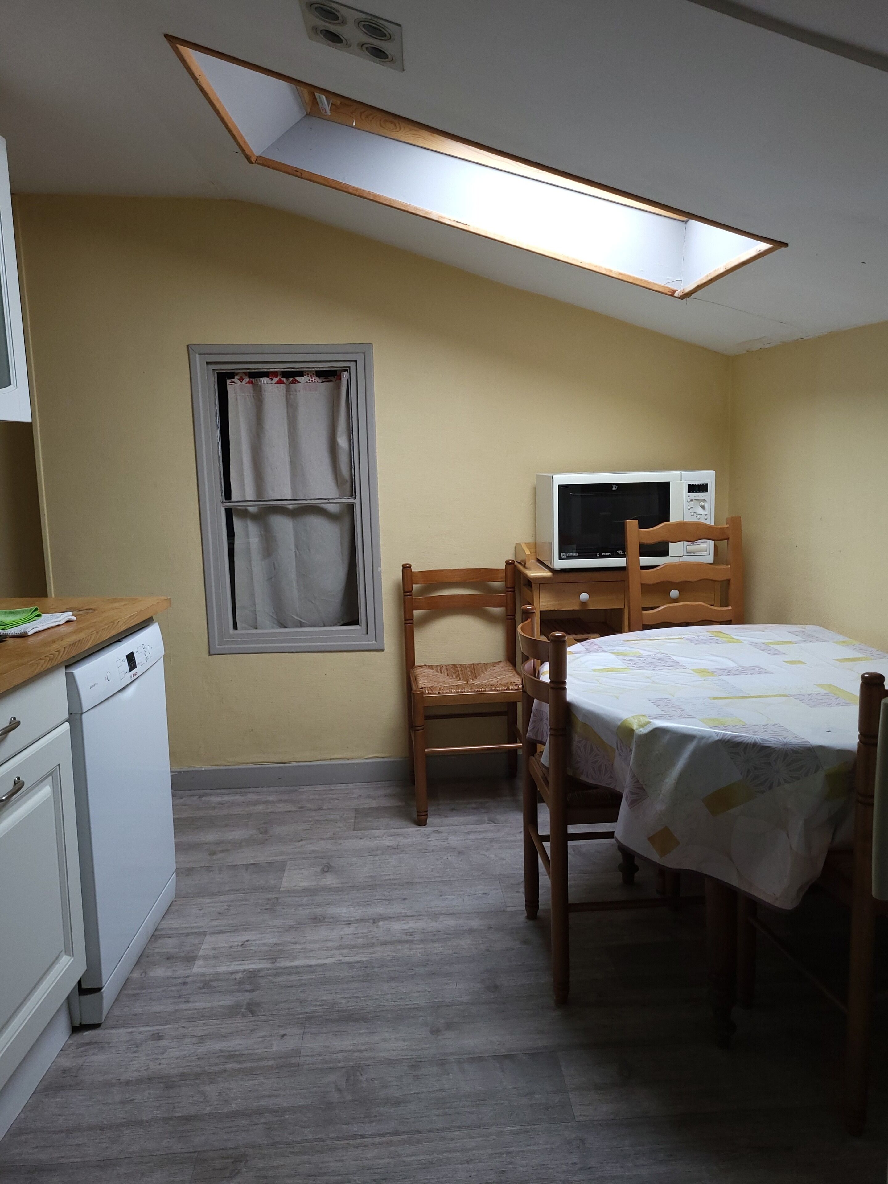 A compact kitchen with a microwave, wooden chairs, a table with a tablecloth, and a window with curtains.