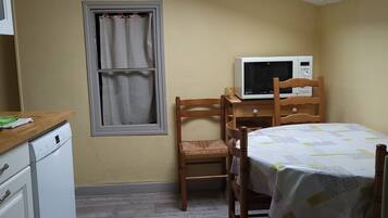 A compact kitchen with a microwave, wooden chairs, a table with a tablecloth, and a window with curtains.