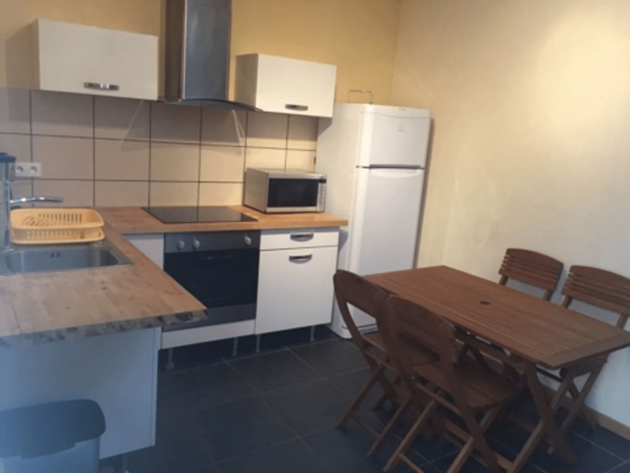 A compact kitchen with a stove, microwave, and refrigerator. There is a wooden table with chairs and a small sink.
