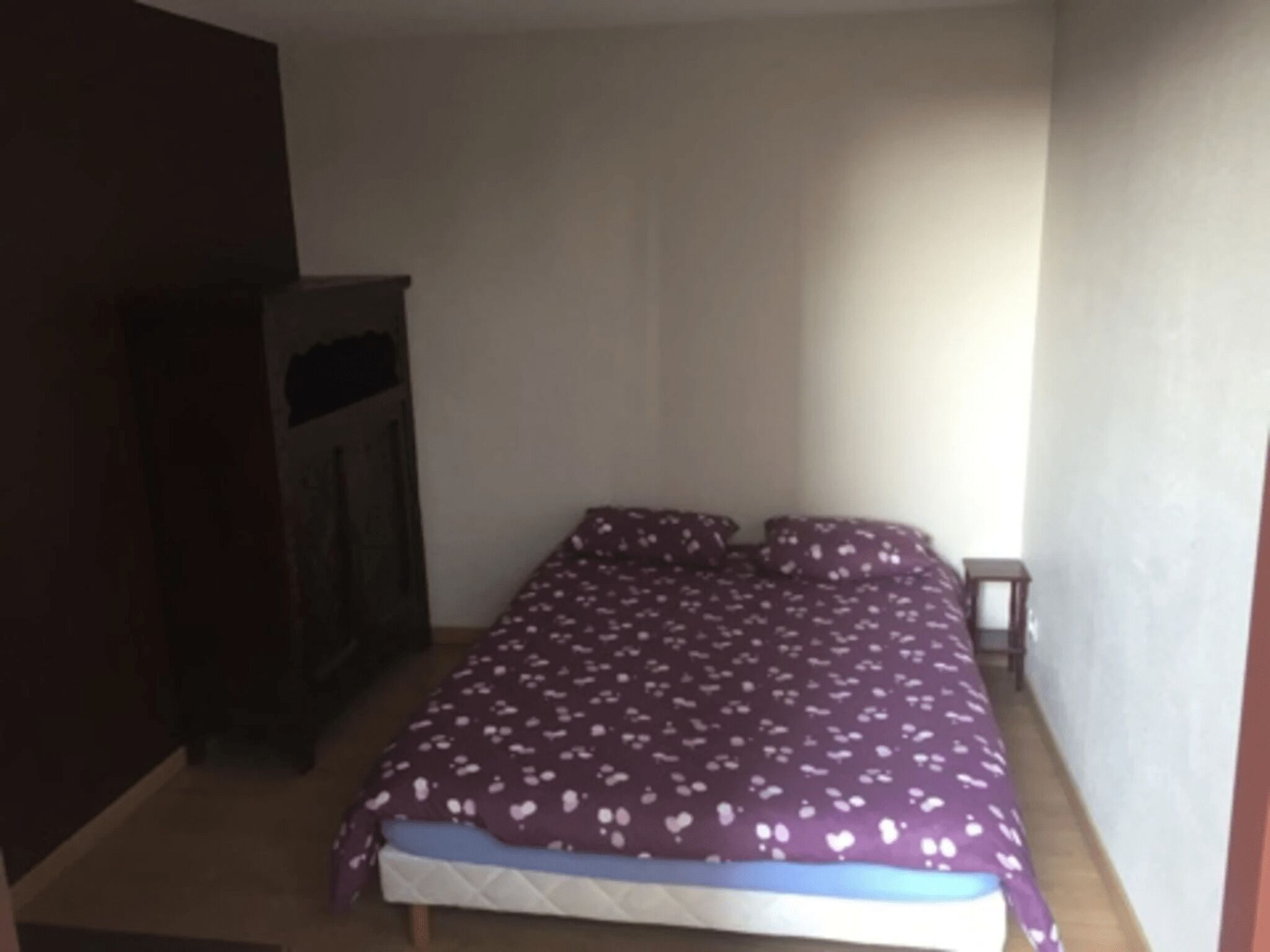 A small, dimly lit room with a single bed, a wooden wardrobe, and a small table.