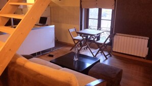 A cozy room with a sofa, a small table, and a chair. There is a wooden staircase leading to another level. A window with a view of the outside is visible. - DUPLEX UNDER THE SLOPES ON THE BANKS OF THE LOIRE (APT N°22) (Roanne)