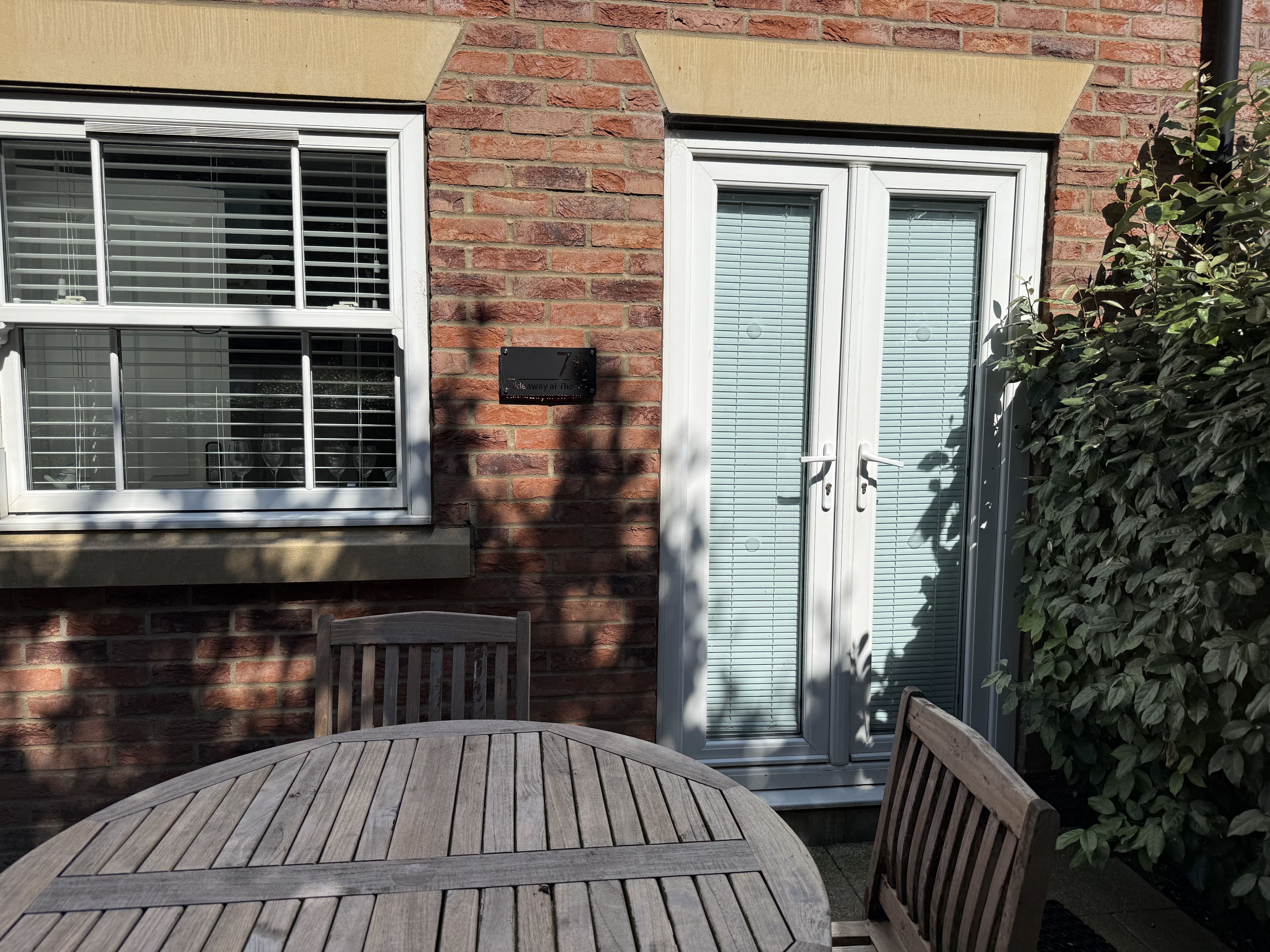 A brick house with a patio table and chairs, a window with blinds, and a door with a mailbox.