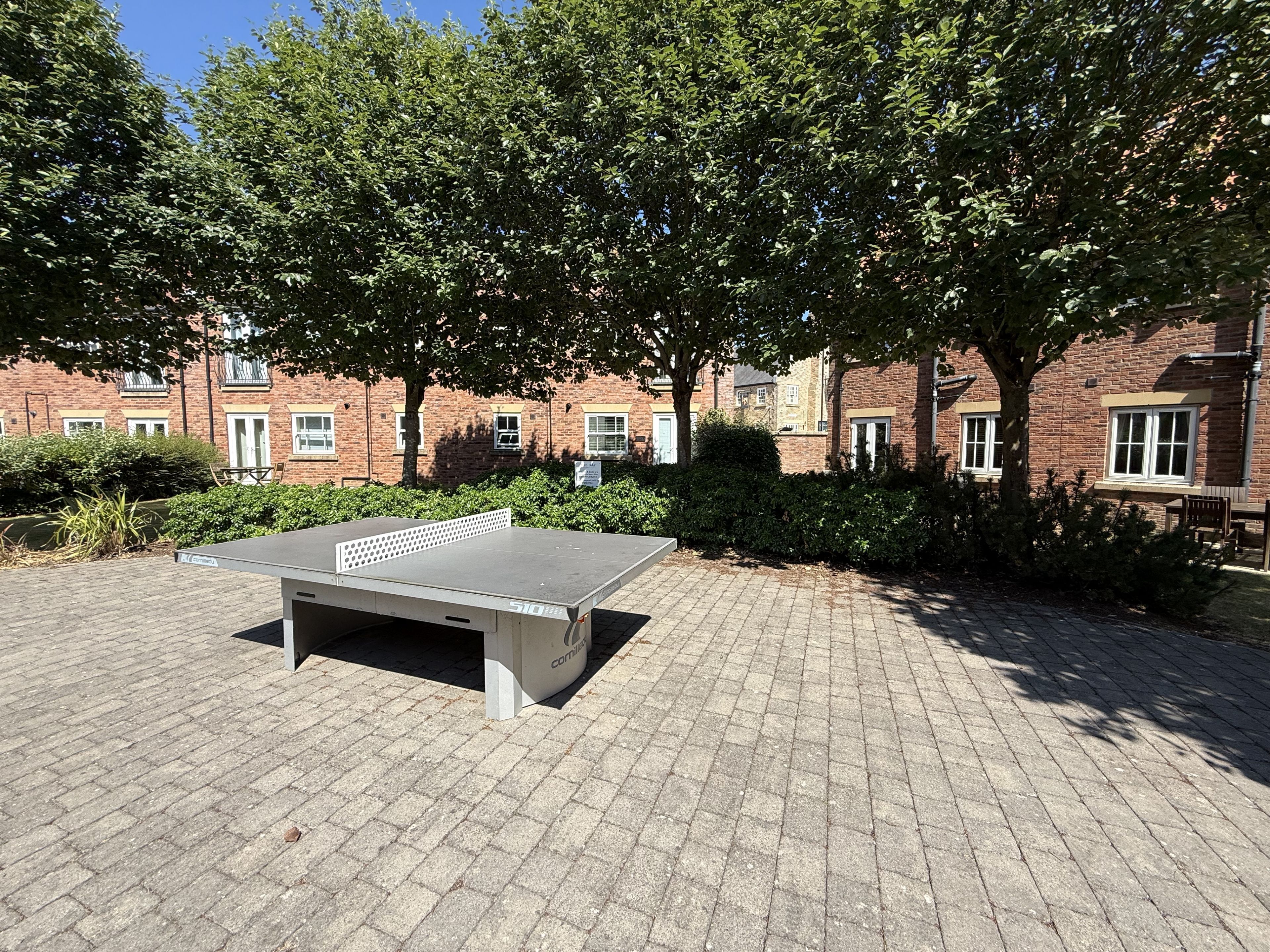 Une table de ping-pong installée dans un espace extérieur, avec des bâtiments en briques et des arbres en arrière-plan.