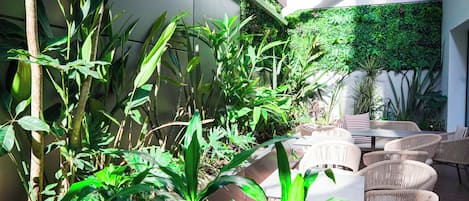 Un espacio patio con mobiliario de mimbre y abundante follaje, incluyendo plantas altas y un muro jardín vertical.