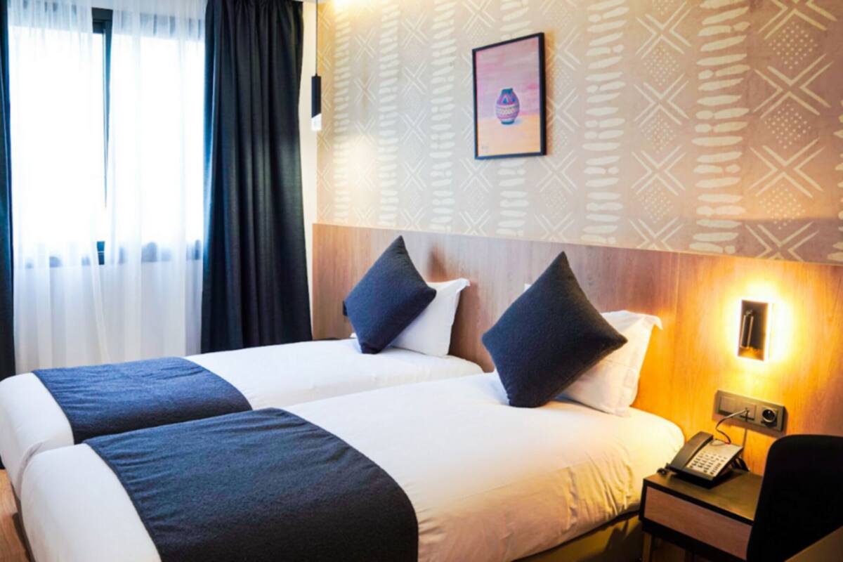 A hotel room with two beds, a bedside table with a phone, and a wall-mounted light.