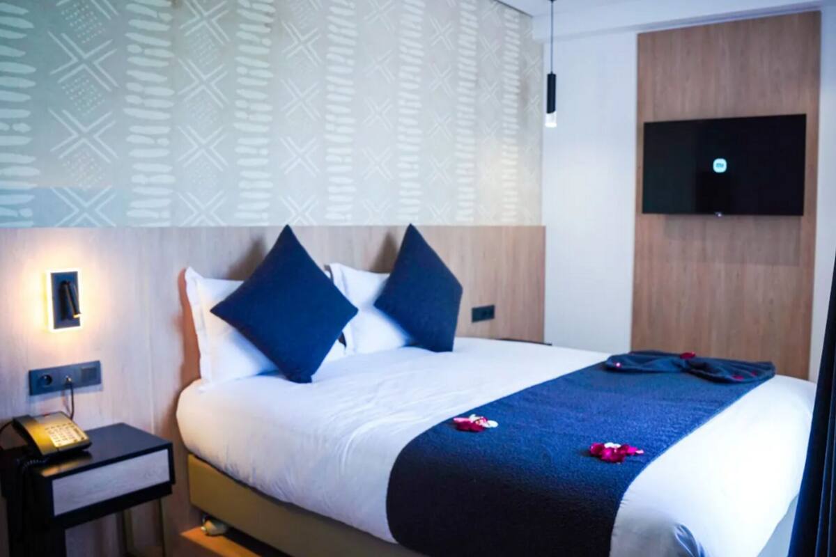 A hotel room with a bed, a bedside table with a telephone, a wall-mounted TV, and a patterned wall.