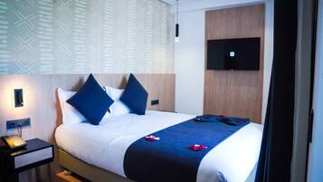 A hotel room with a bed, a bedside table with a telephone, a wall-mounted TV, and a patterned wall.