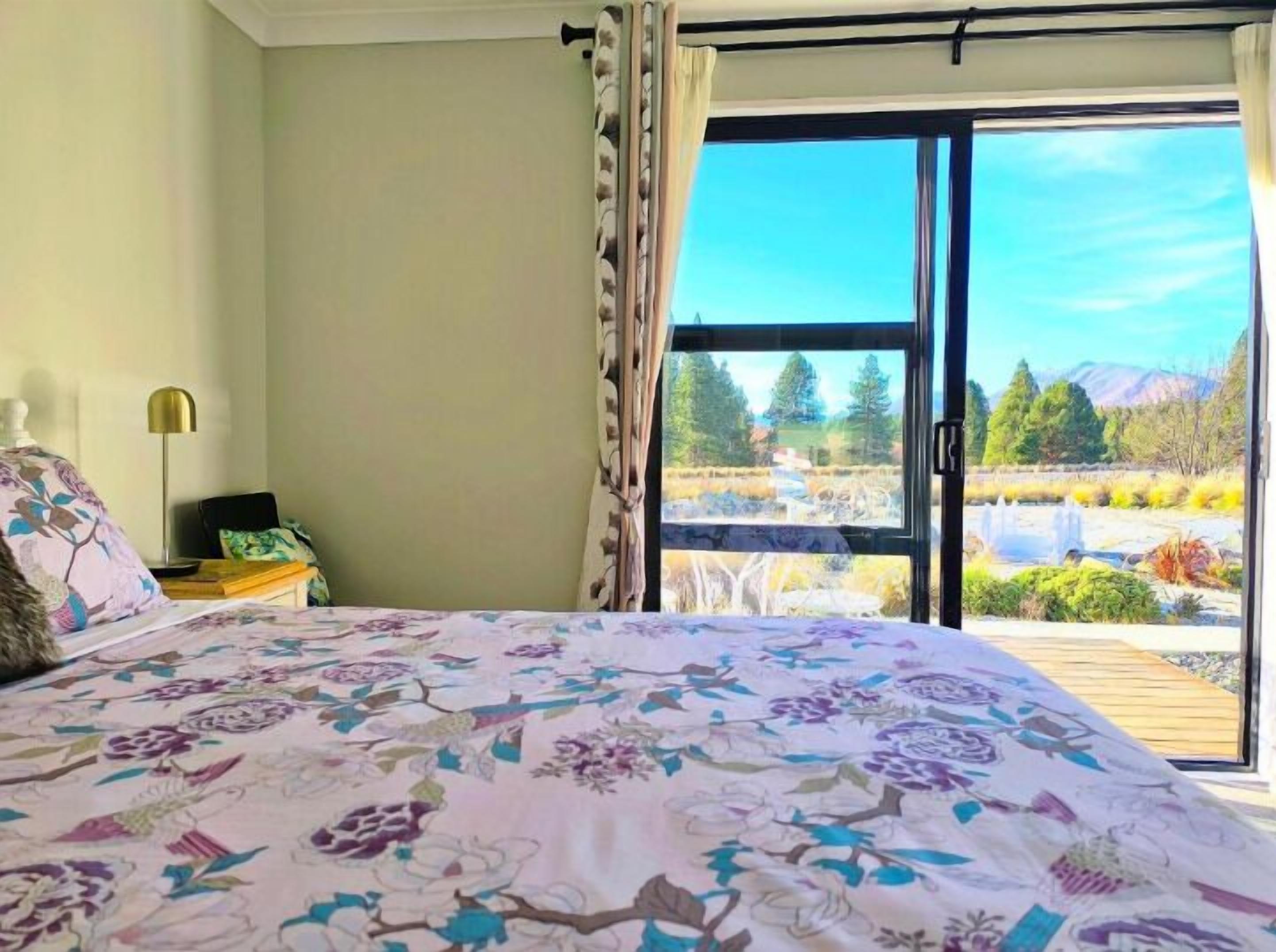 Camera da letto con un letto, un comodino, una lampada e vista su un paesaggio naturale attraverso una porta-finestra in vetro.
