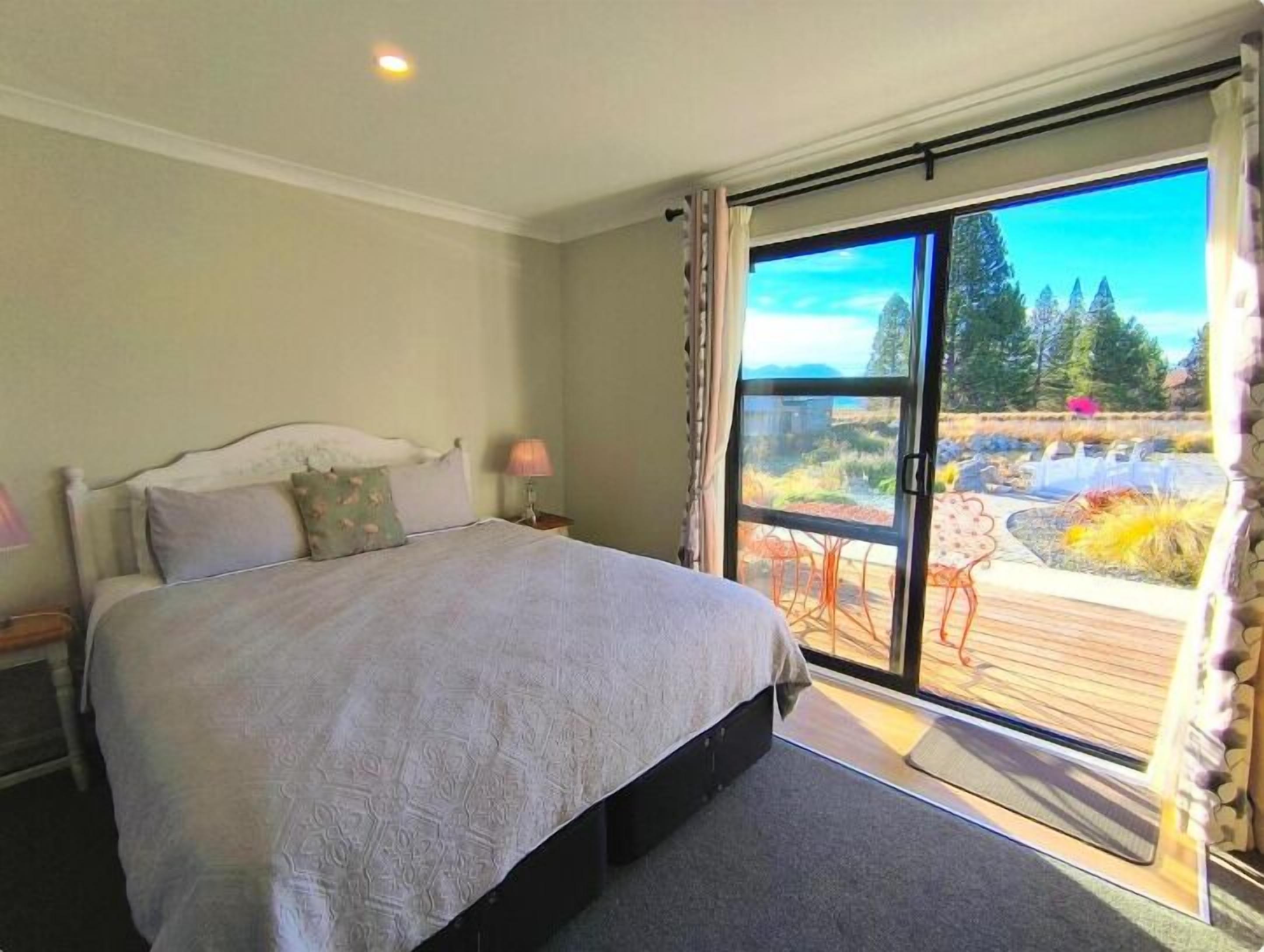A bedroom with a large bed, a nightstand, a lamp, and a view of a garden through a sliding glass door.