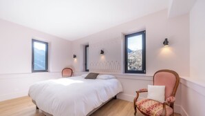 Ein Schlafzimmer mit einem Bett, einem Sessel und zwei Fenstern mit Wandleuchten.