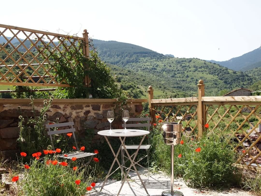 A patio with a table and chairs, a wine glass, and a metal container, surrounded by red poppies and a stone wall.