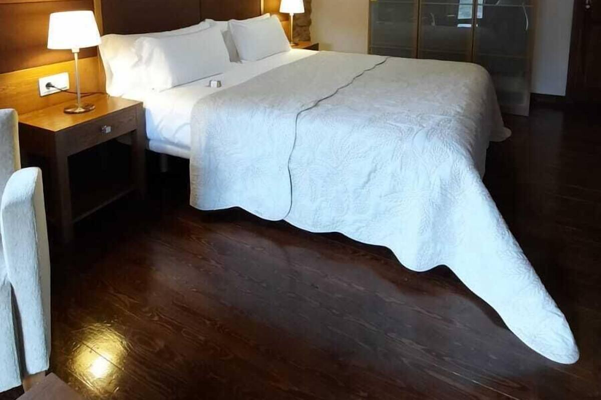 Un dormitorio con techo de madera, pared de piedra, cama con ropa de cama blanca, dos lámparas de noche y un armario con puertas de vidrio.