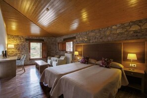 Quarto de hotel com duas camas, parede de pedra, teto de madeira e uma escrivaninha com cadeira.